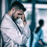 Physician holding his head against a wall with another physician behind him