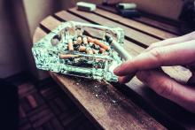 Man smoking a cigarette holding a burning filter tip in his hand alongside a glass ashtray full of ash and dead butts.