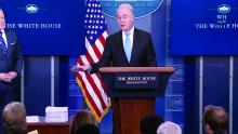 Dr. Price stands at a podium in the White House briefing room. A flag and two large monitors are in the background.